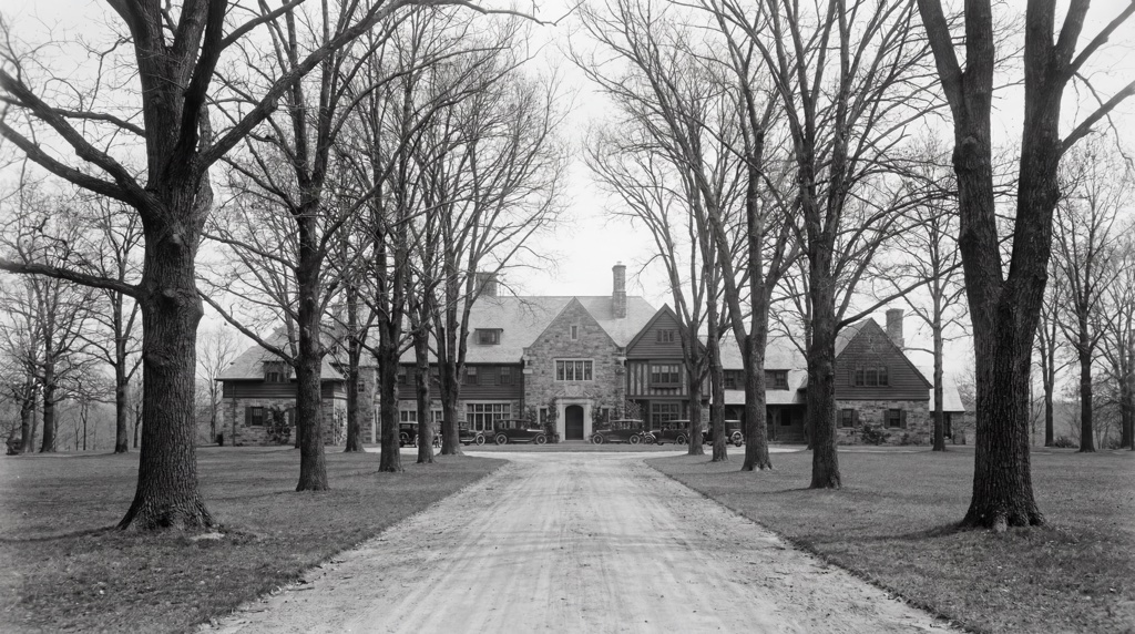 Historic B&W of the manor — large stone house framed by tall trees, vintage cars in the drive.